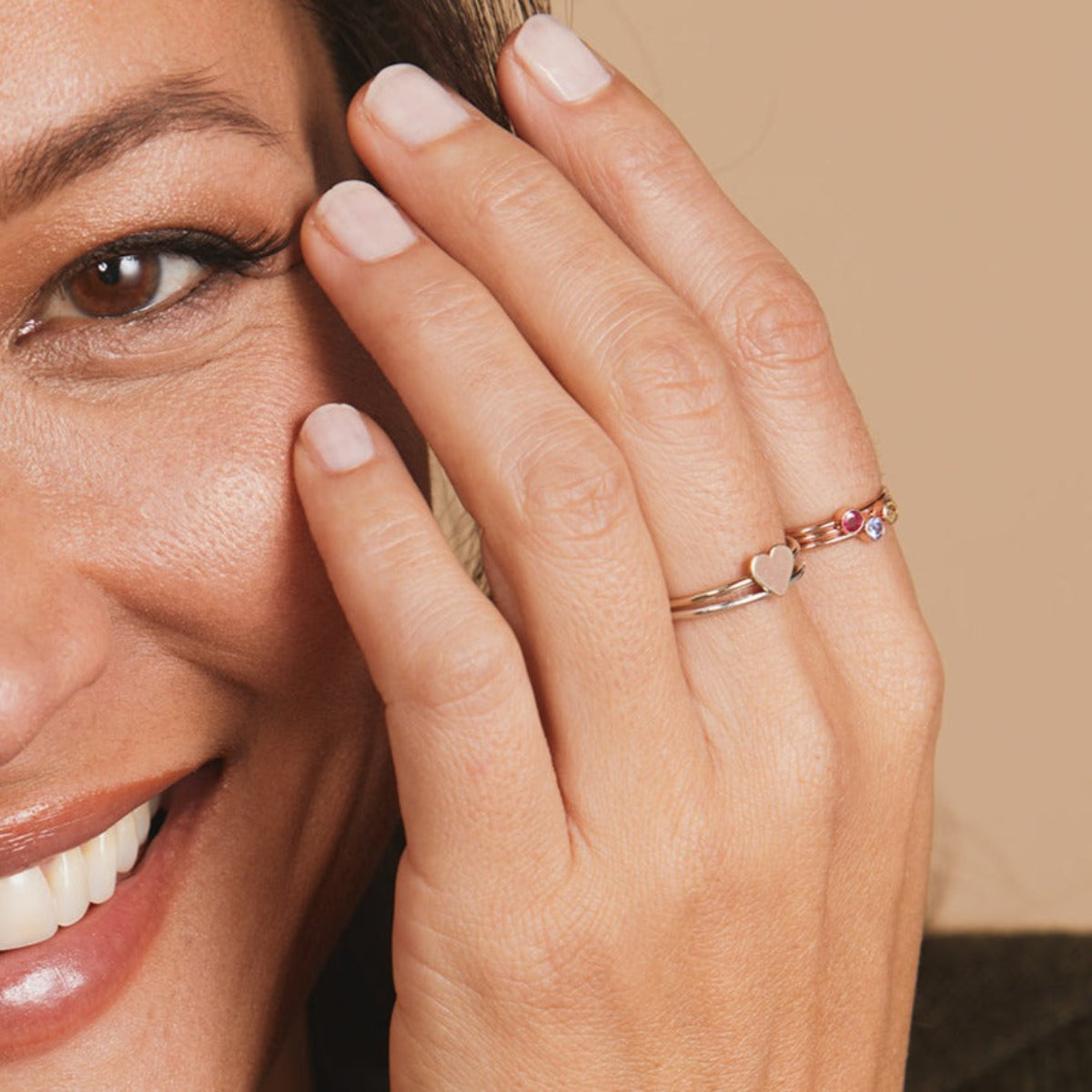 Close-up of a woman's hand wearing a white gold Heartfelt Ring and three Birthstone Rings, against light neutral background. 