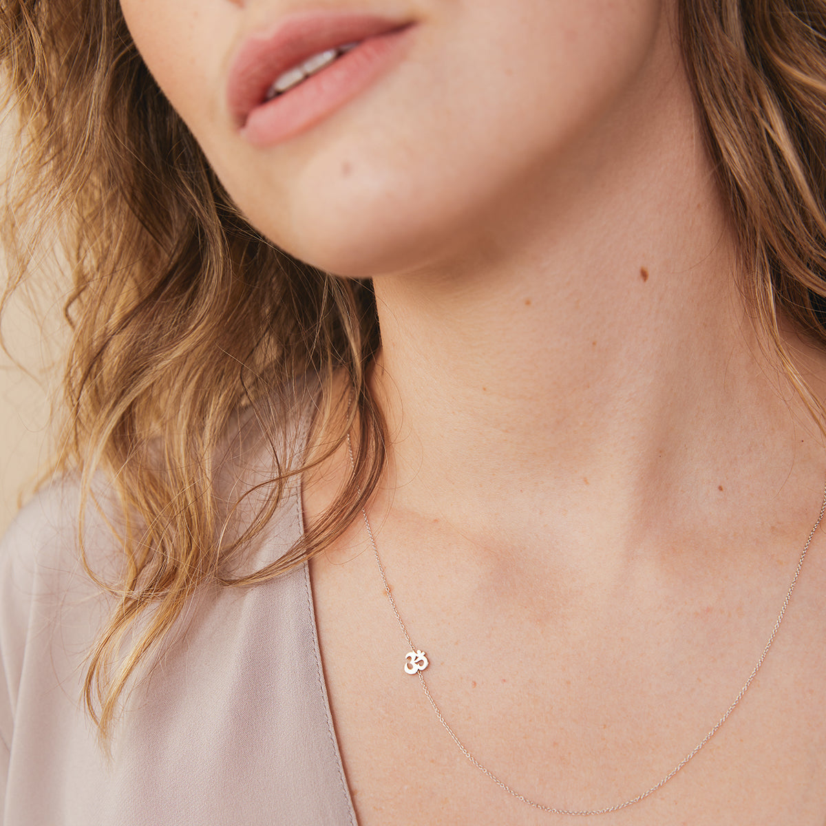 A close-up of a woman's neck wearing a  white gold 14k Gold Asymmetrical Charm Necklace - Om with a cable chain, styled with a neutral top. 