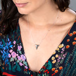 Close-up of a brunette woman's décolletage wearing a white gold Sharktooth Necklace with a navy top featuring flowers in various colours against a grey background. 