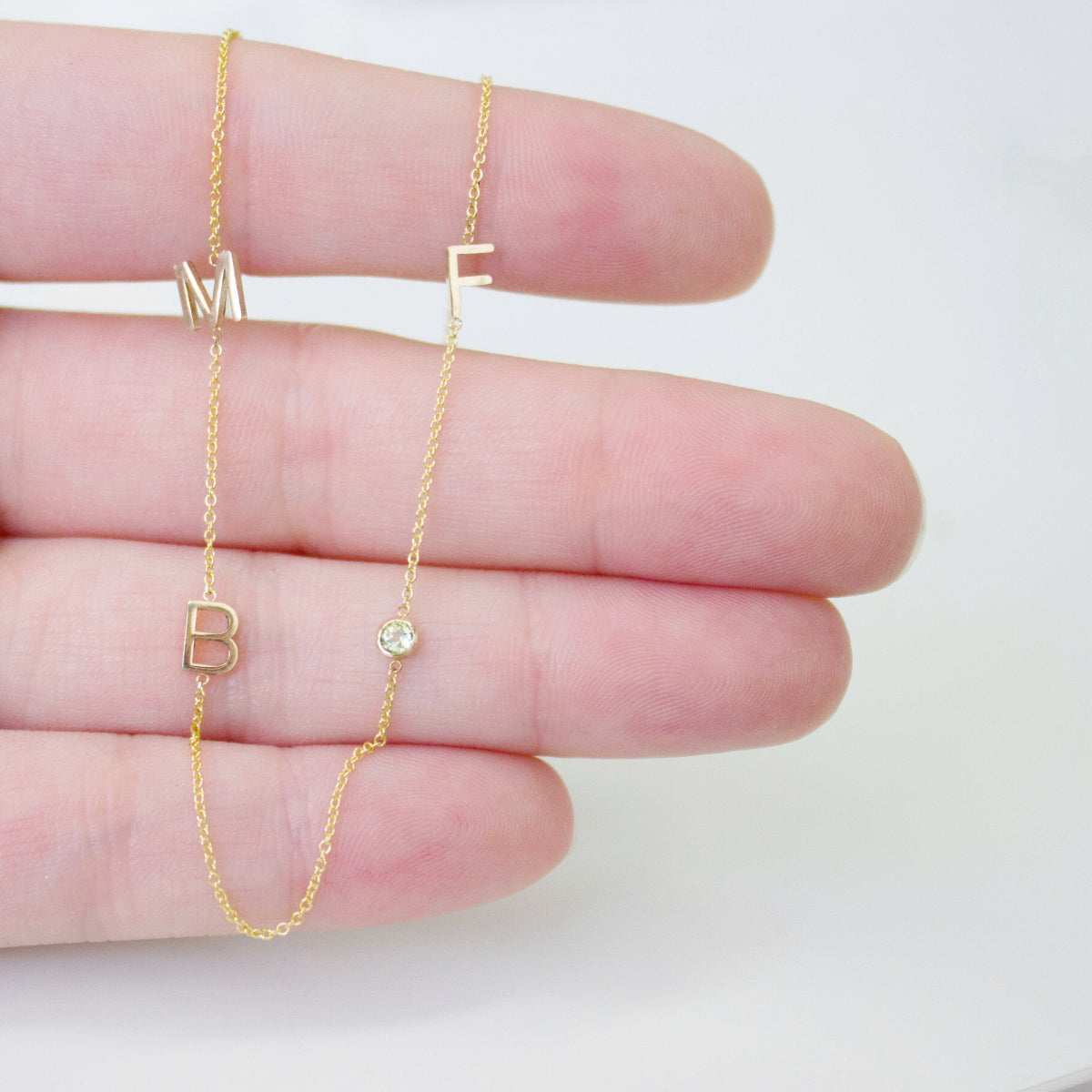 A Custom Gold Necklace in yellow gold with three letter charms, M, F, and B, and a birthstone, held between fingers on a white background.