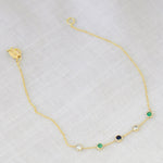 A Custom Gold Bracelet – 5 Letters with five birthstones (two diamond, one sapphire and two emerald) on a white fabric background