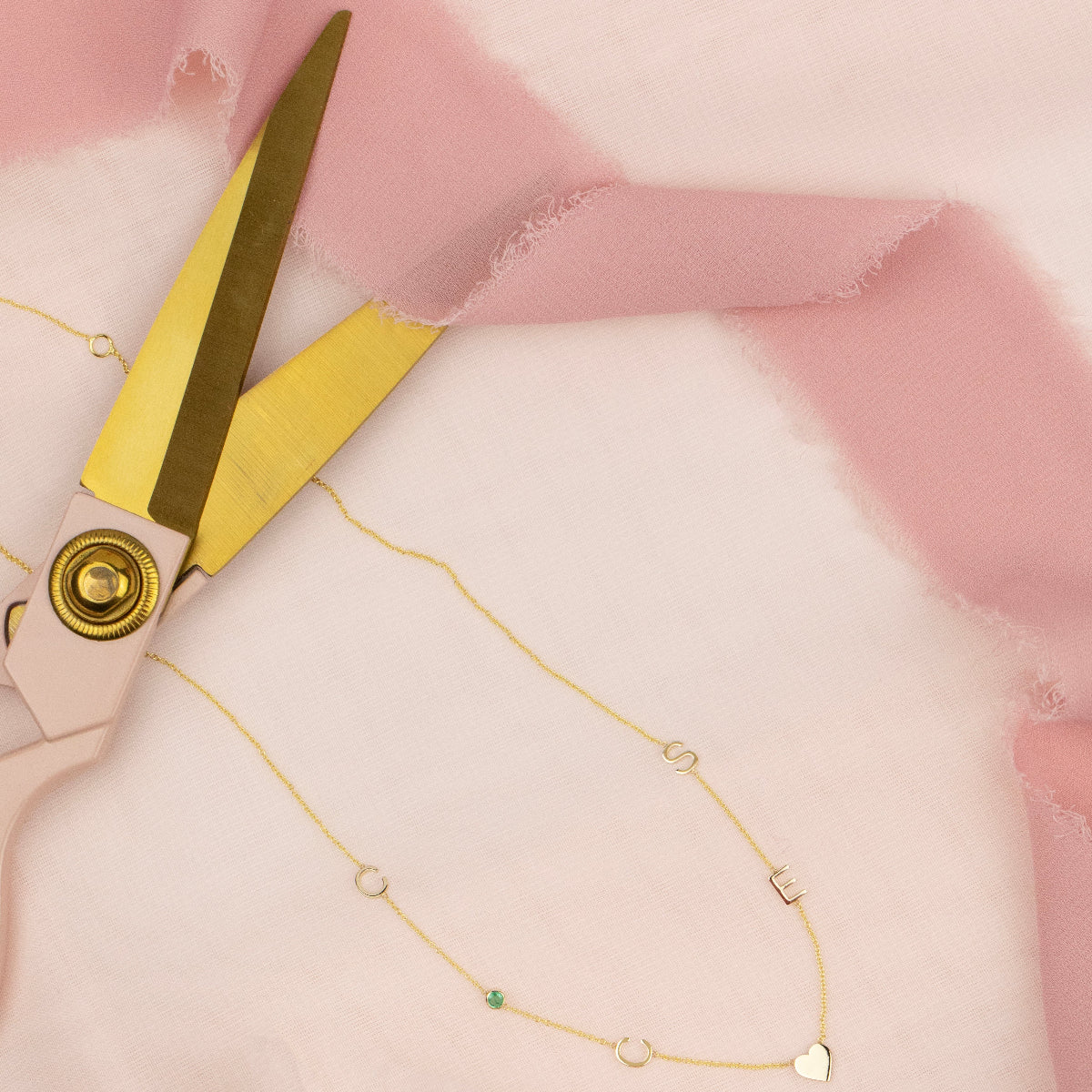 A Custom Gold Necklace in yellow gold with 6 charms (two Cs, S, E, a birthstone and a heart) on pink fabric with a pair of gold scissors.