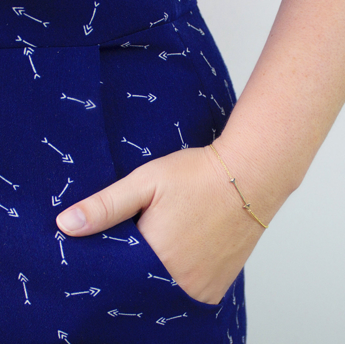 A hand wearing a Straight as an Arrow bracelet in yellow gold, in the pocket of navy blue pants with a white arrow pattern. 
