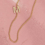 A yellow gold beaded necklace with a pave letter M charm on a pink fabric background. 