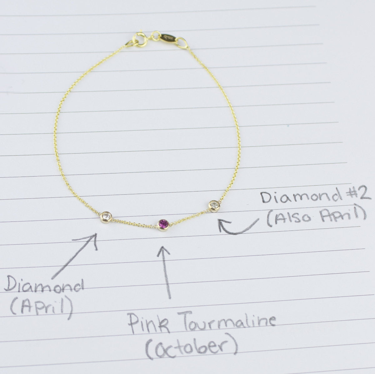 A yellow gold Custom Gold Bracelet with 3 letters and three birthstones (two diamonds and a pink tourmaline) displayed on a sheet of paper with annotations listing the birthstones and their corresponding months.