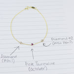 A yellow gold Custom Gold Bracelet with 3 letters and three birthstones (two diamonds and a pink tourmaline) displayed on a sheet of paper with annotations listing the birthstones and their corresponding months.