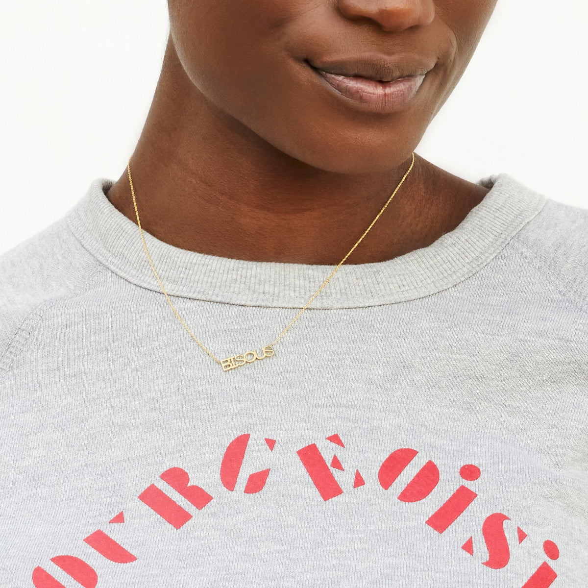 A close-up of a woman wearing a BISOUS Necklace with a light grey sweater with red writing. 