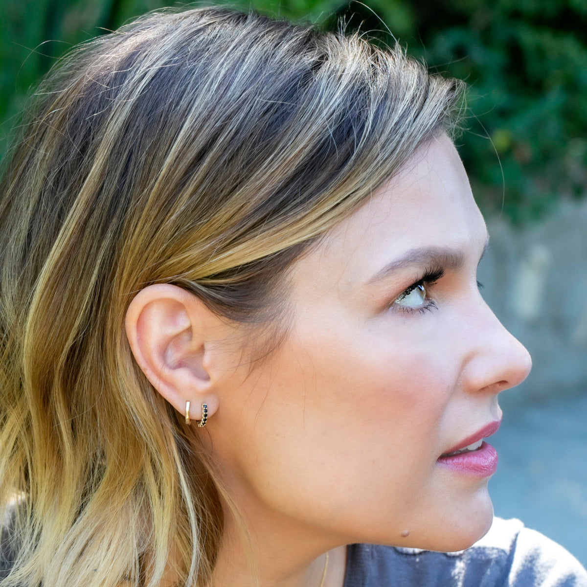 A woman wearing an Alli Webb Faceted Huggie and Onyx Huggie with blonde hair against a blurred greenery background. 