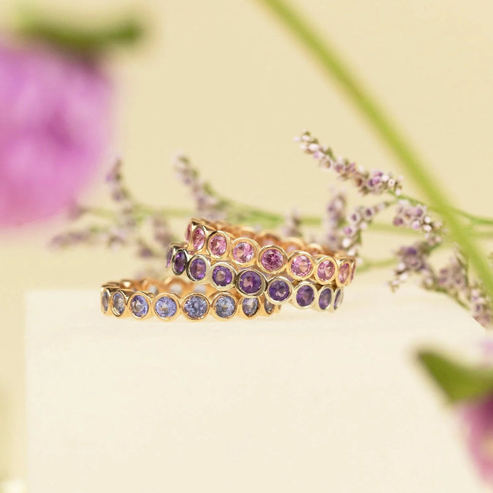 The Peony Bouquet Ring, The Violet Bouquet Ring and The Periwinkle Bouquet Ring against a light yellow blurred background with purple flowers. 