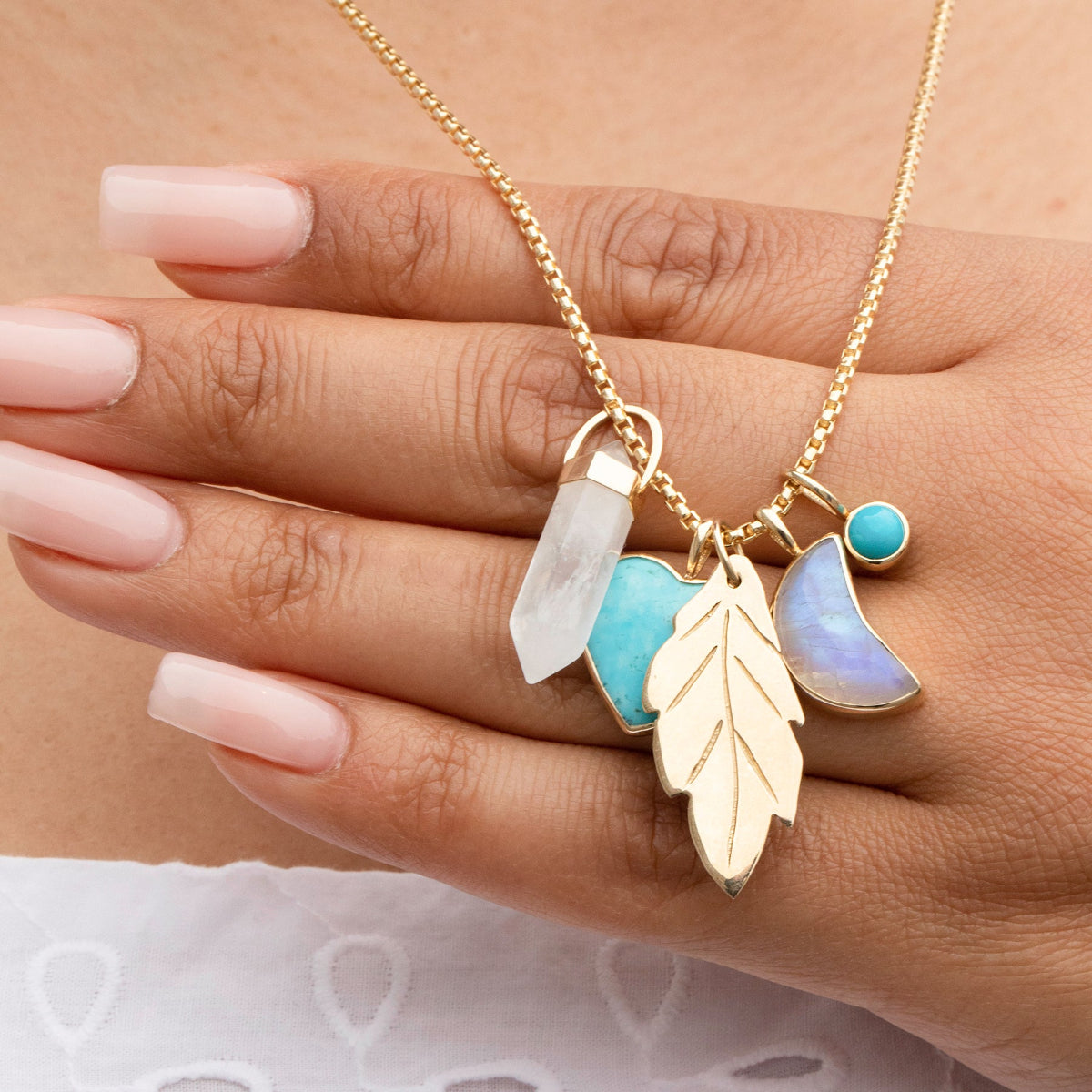 A yellow gold Box Chain with several charms, including a Balance Crystal Charm, Turquoise Heart Charm, Moonstone Crescent Charm, 20th Anniversary Leaf Pendant and a Turquoise Dangle Charm held by a hand in the background. 