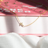 A 14k Gold Asymmetrical Charm Necklace - Cross in yellow gold and cable chain, laid on top of a book with a dark pink cover on a white fabric surface.