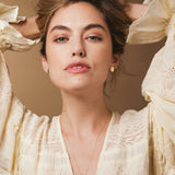 A woman wearing The Clarity Retreat Necklace and Solid Eclipse Earrings - Small, styled with a light yellow long sleeve top against a light brown background. 