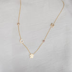 A Custom Gold Necklace in yellow gold with five charms, two birthstones, the letters C and M, and a paw, on a neutral background.
