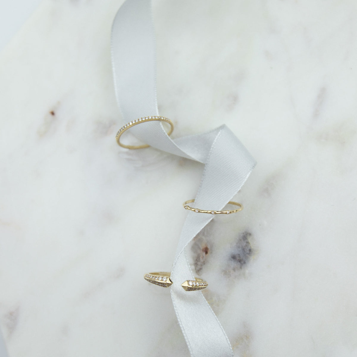 A Pave Diamond Ring, 4 Diamond Ring and Open Diamond Ring on white ribbon against a white marble background.