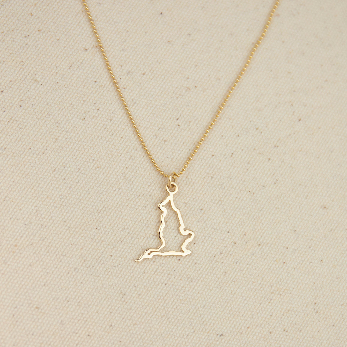 An England pendant necklace in yellow gold with a beaded chain on a beige fabric background. 