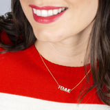 A close-up of a woman smiling and wearing a FEMME Necklace with a red and white knit jumper, with her brown hair visible. 