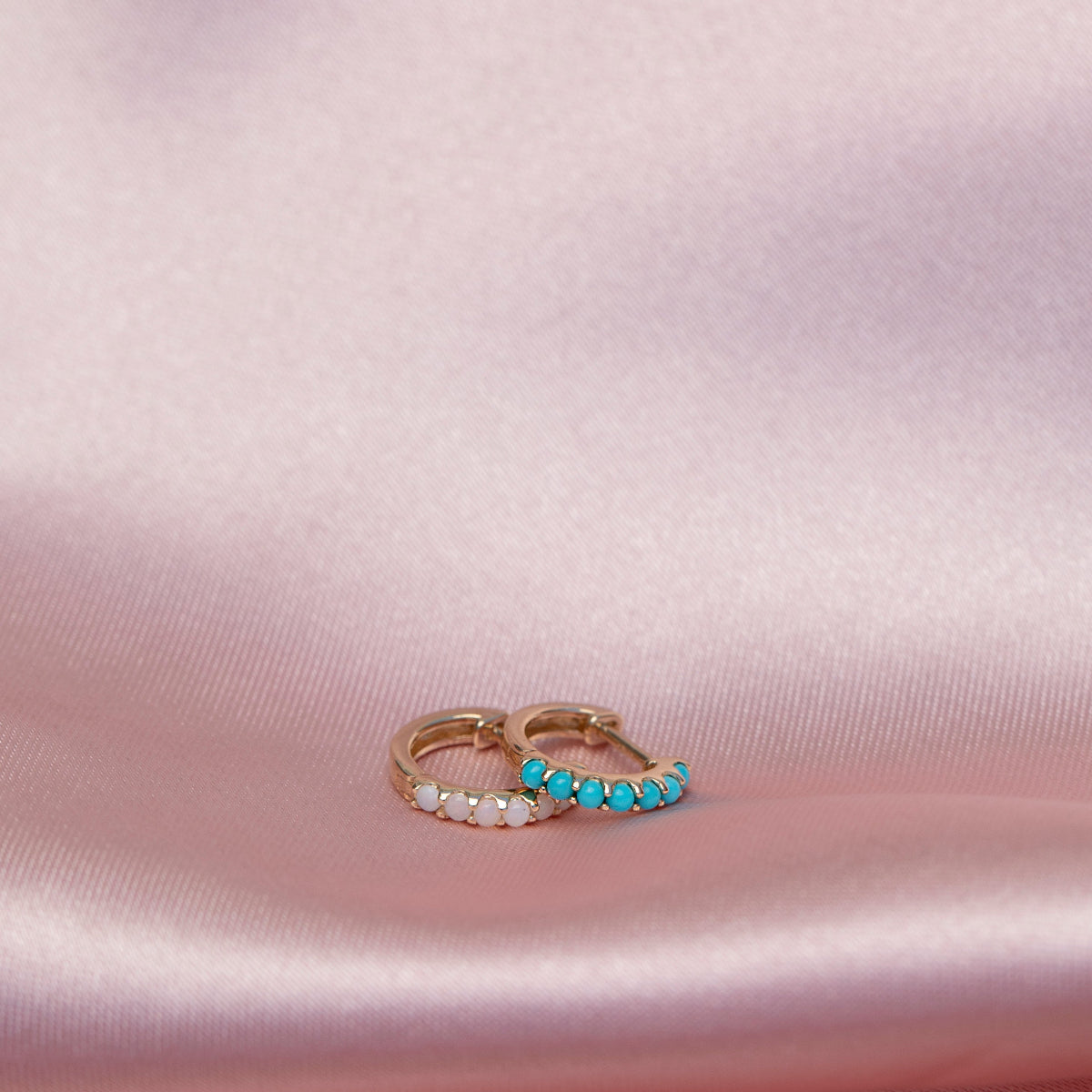 An Alli Webb Opal Huggie and Turquoise Huggie on a light pink silk fabric. 