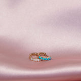 An Alli Webb Opal Huggie and Turquoise Huggie on a light pink silk fabric. 