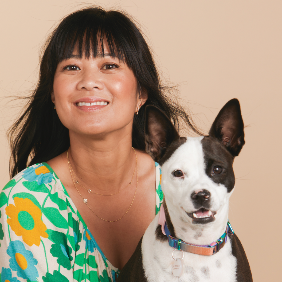 A woman wearing a yellow gold 14k Gold Asymmetrical Charm Necklace - Paw and a Custom Gold Necklace - 2 Letters, wearing a green, blue and yellow floral top, holding a dog against a beige background.