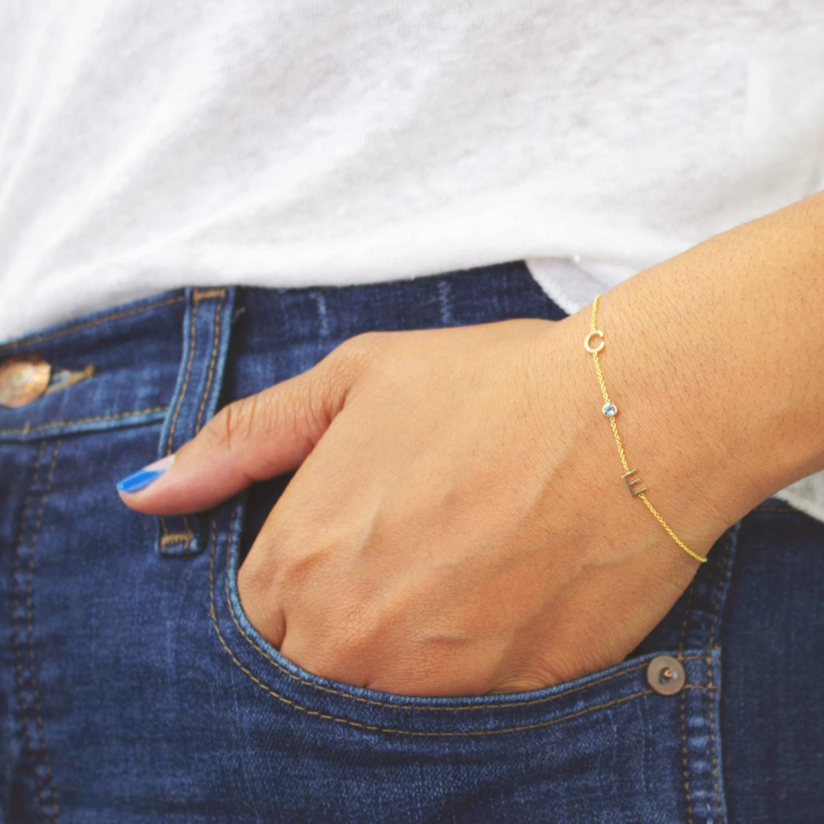 A hand wearing a yellow gold Custom Gold Bracelet with 3 letters (two letters C and E and an aquamarine birthstone), in a dark blue denim jean pocket, styled with a white top.