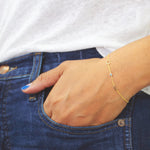 A hand wearing a yellow gold Custom Gold Bracelet with 3 letters (two letters C and E and an aquamarine birthstone), in a dark blue denim jean pocket, styled with a white top.