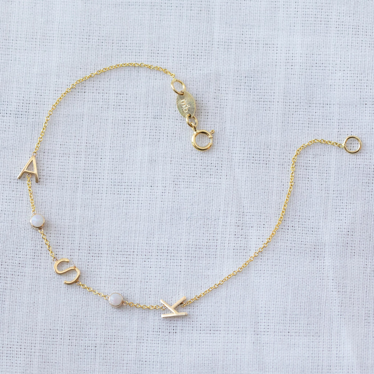 A yellow gold Custom Gold Bracelet - 5 letters, with the letter charms A, S and K and two birthstones on a white fabric background. 