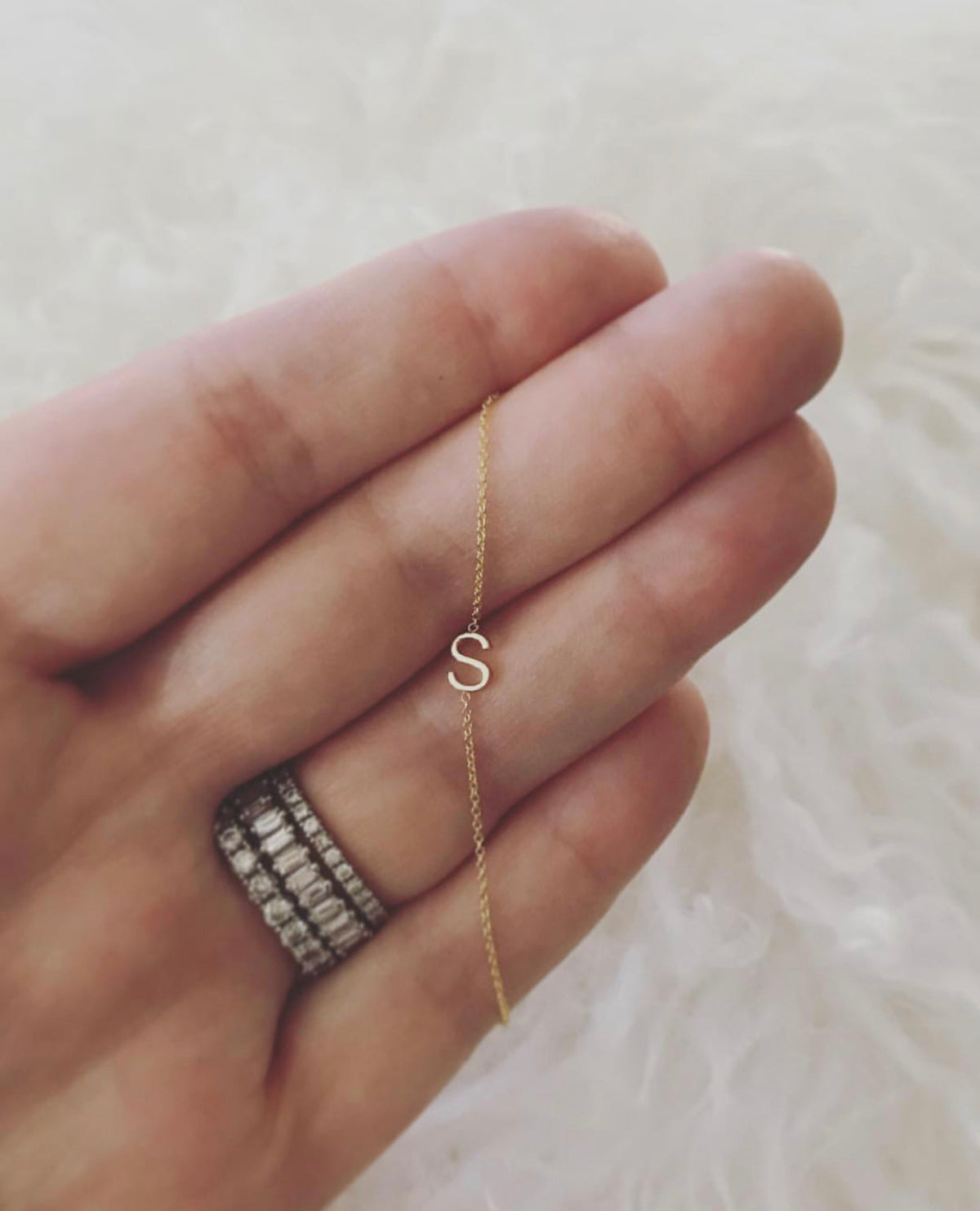 A close-up of a hand wearing three rings, with a yellow gold necklace featuring a letter S charm wrapped around the fingers, set against a cream furry background.