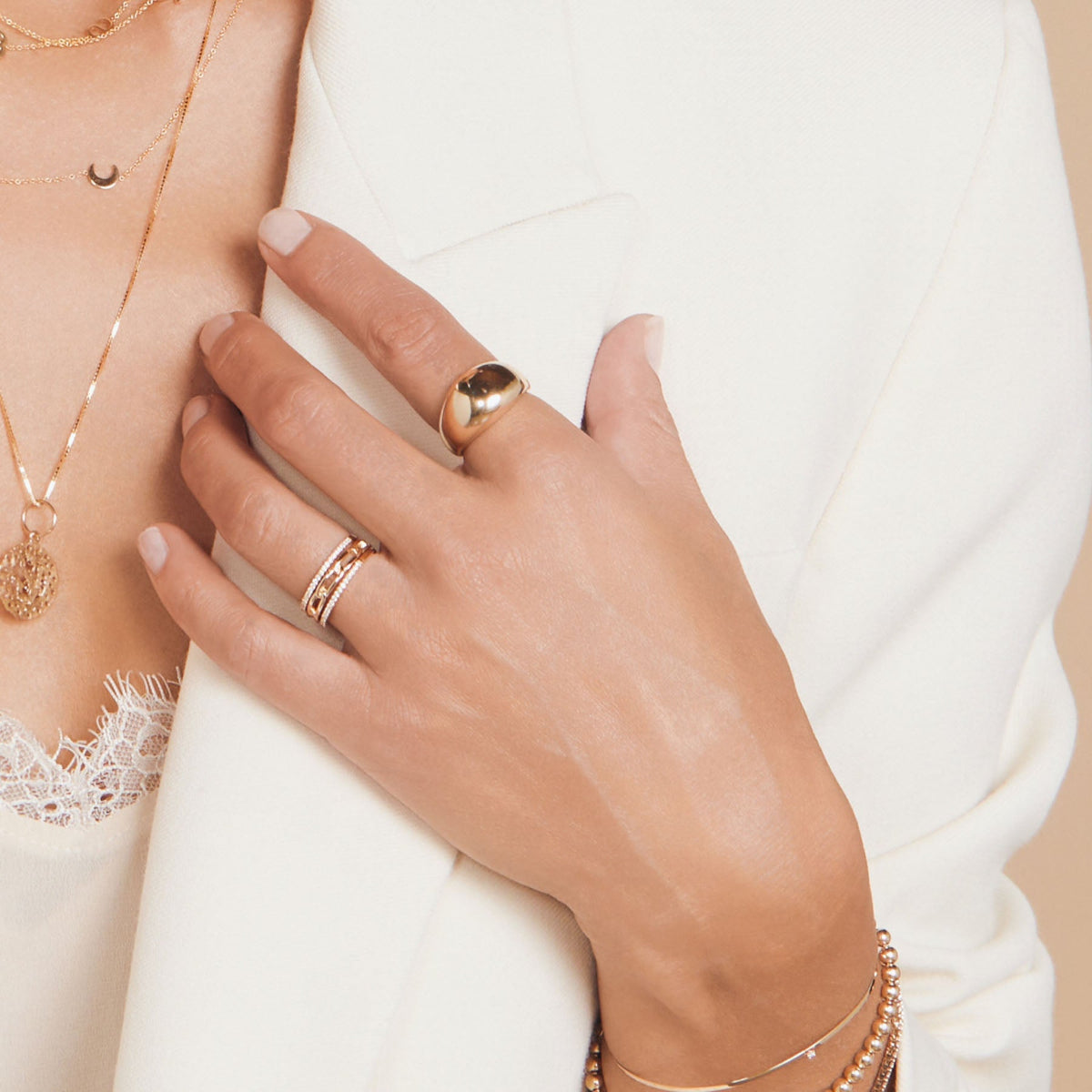 Close-up of a woman’s hand wearing a Link Ring, two Pavé Diamond Rings, a Paris Ring, an Off the Cuff Bracelet, and a Bubble Bracelet, styled with a 14k gold Asymmetrical Charm Necklace (Moon) and other yellow gold necklaces, paired with a white blazer and lace top.