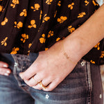 Close-up of a person arm and hand wearing a Love Bracelet and gold rings, including a Geo Stacking Ring, Pave Geo Ring with a black and yellow floral-patterned shirt and blue jeans. 