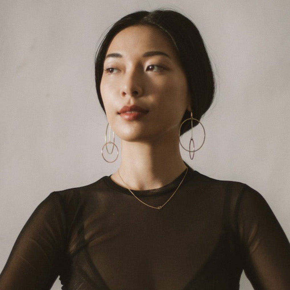 A woman in a black sheer long sleeve top wearing a yellow gold necklace with a letter S charm and drop earrings with circles. 