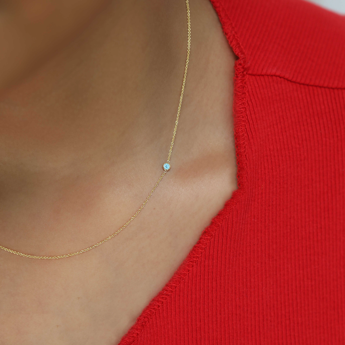 A close-up of a neck wearing a yellow gold 14K Gold Asymmetrical Birthstone Necklace - Aquamarine (March), styled with a red top. 