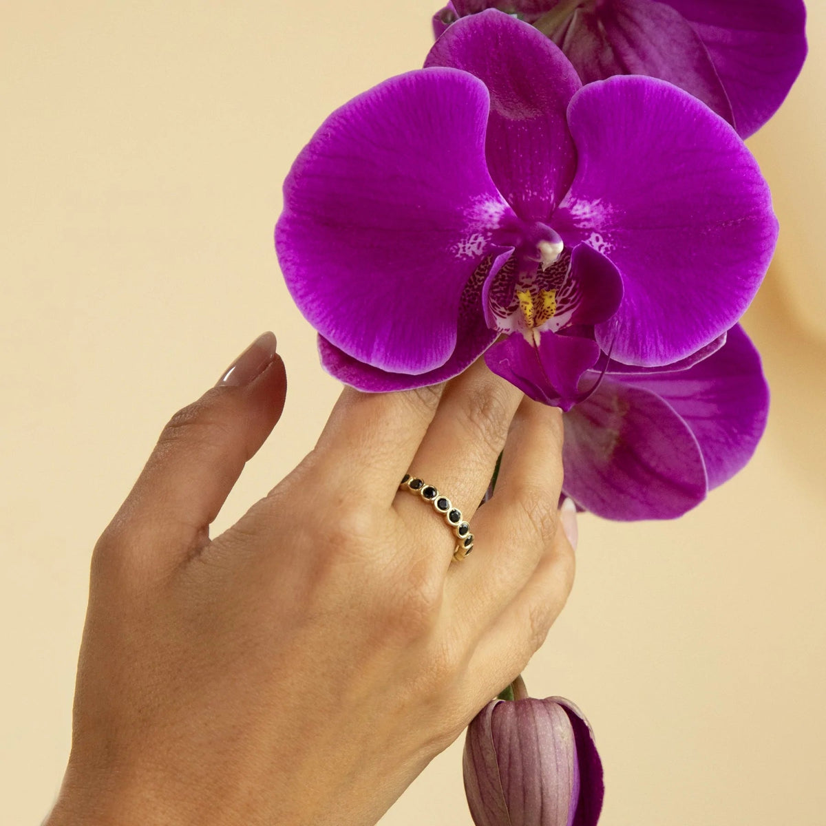 A hand wearing The Orchid Bouquet Ring and holding a purple orchid flower against a light yellow background.