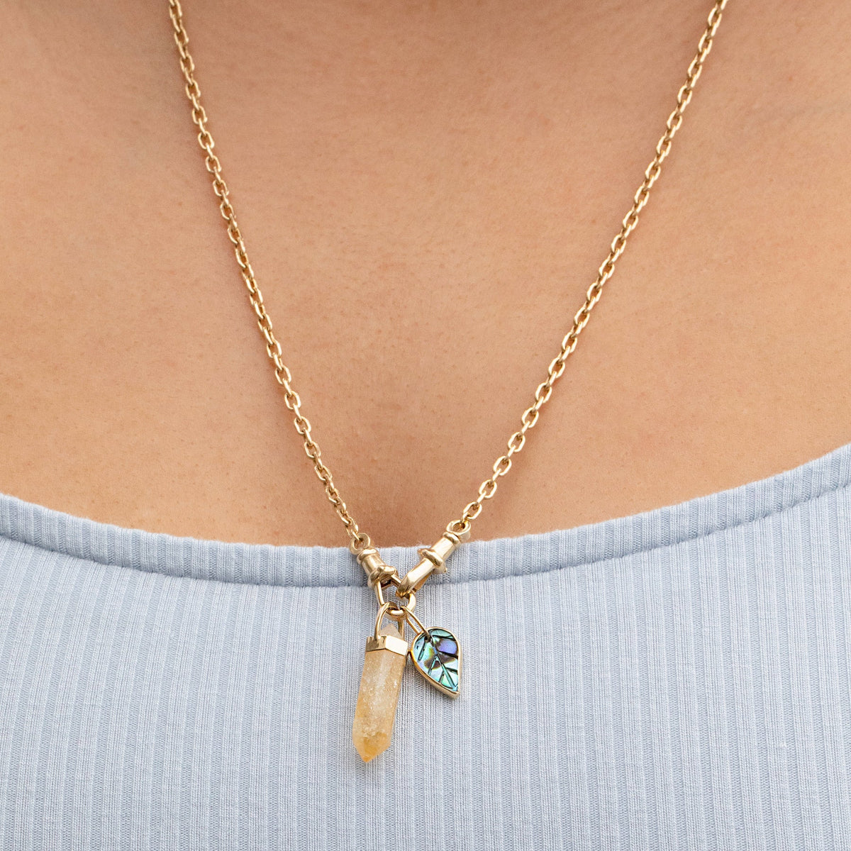 Close-up of a décolletage wearing an Oval Link Chain with an Abalone Leaf Charm and a Balance Crystal Charm with a light blue top. 