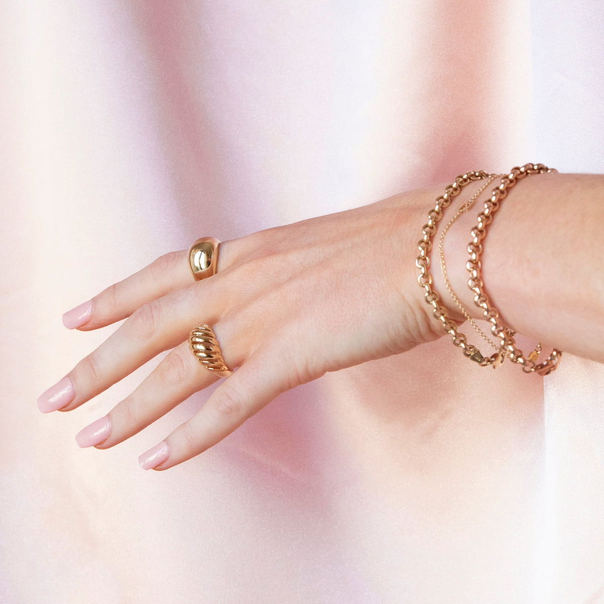 A hand wearing a yellow gold Paris Ring, Etched Paris Ring and two Rolo Bracelets against a blurred background. 