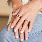 Close-up of hands wearing a yellow gold Paris Ring, two Pavé Diamond Rings, a Link Ring, Trio Stacking Rings, two Bouquet Rings, a Tube Ring, a Rolo Bracelet and a Bubble Bracelet, resting on blue jeans against a blurred background.