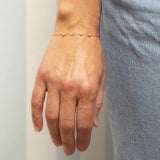 A hand wearing a Custom Gold Bracelet - 4 Letters with four pearl birthstones next to light blue jeans on a neutral stripe background. 