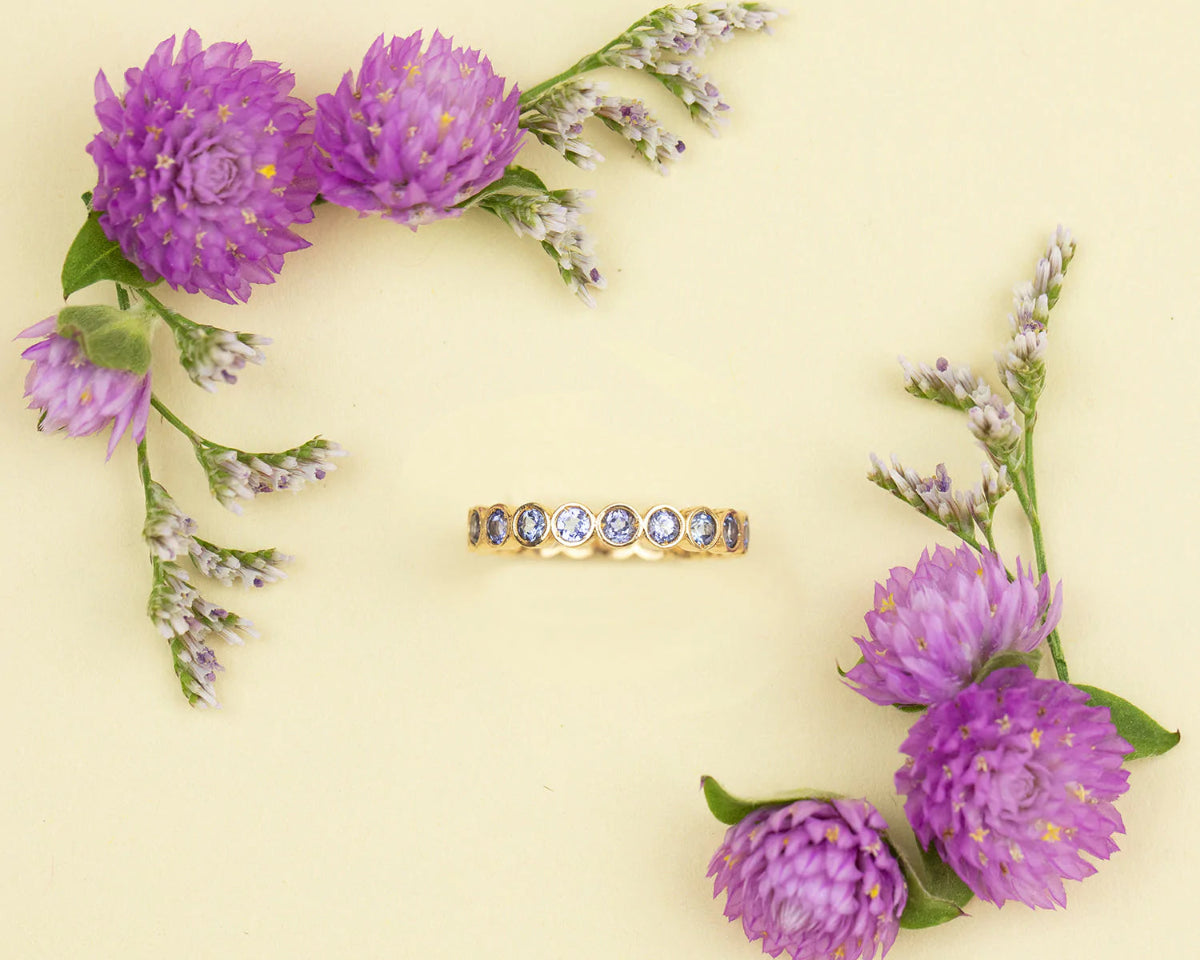 The Periwinkle Bouquet Ring on a light yellow background with purple flowers in two corners. 