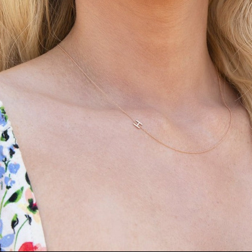A close-up of a woman's neck wearing a gold necklace with a letter H charm. 