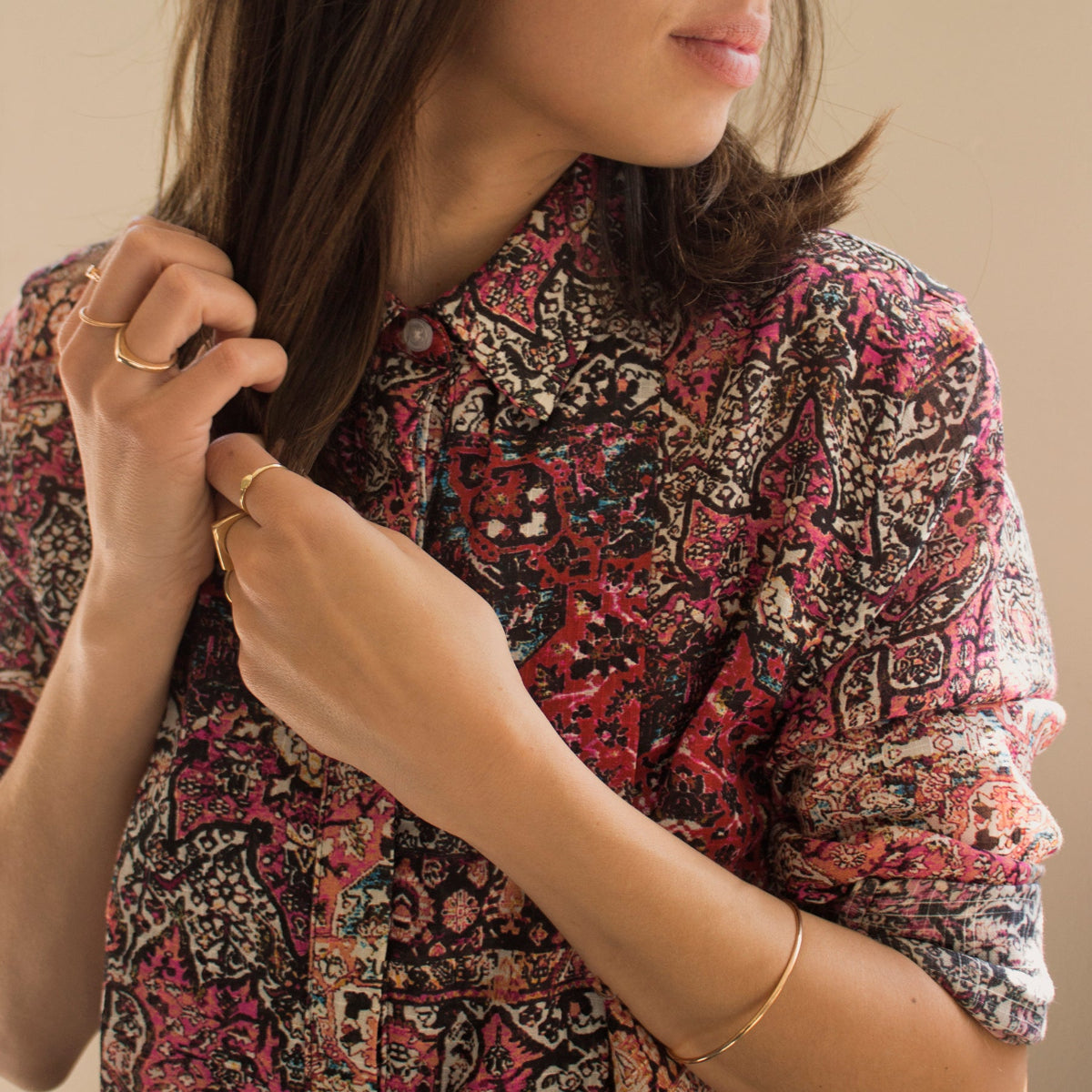 A woman wearing several yellow gold rings, including a Petal Ring, Geo Stacking Ring Triangle and Geo Stacking Ring Square, along with a Flirt Bangle, styled in a colorful patterned top against a light neutral background. 