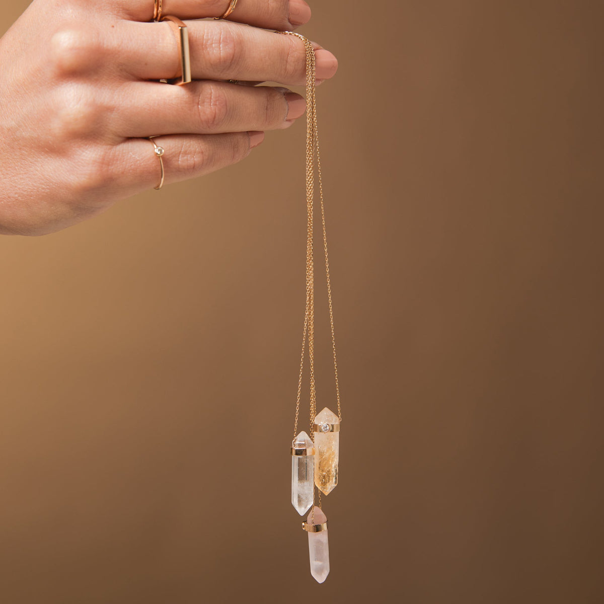 A hand holding three Retreat Necklaces in Clarity, Happiness and Love, a Geo Stacking Ring Square and a Birthstone Ring on a light brown background. 