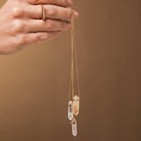 A hand holding three Retreat Necklaces in Clarity, Happiness and Love, a Geo Stacking Ring Square and a Birthstone Ring on a light brown background. 