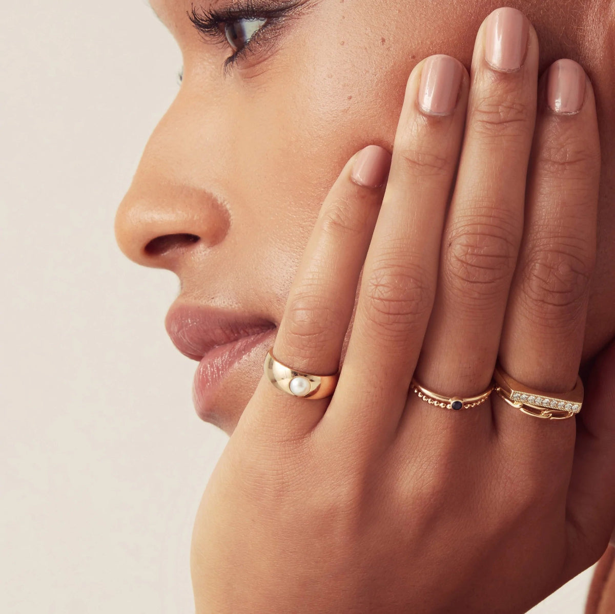 Close-up of a woman wearing a Perla Pinky Ring, Birthstone Ring, Delicate Beaded Band, Link Ring and Pave Geo Ring looking to the side against a light grey background. 