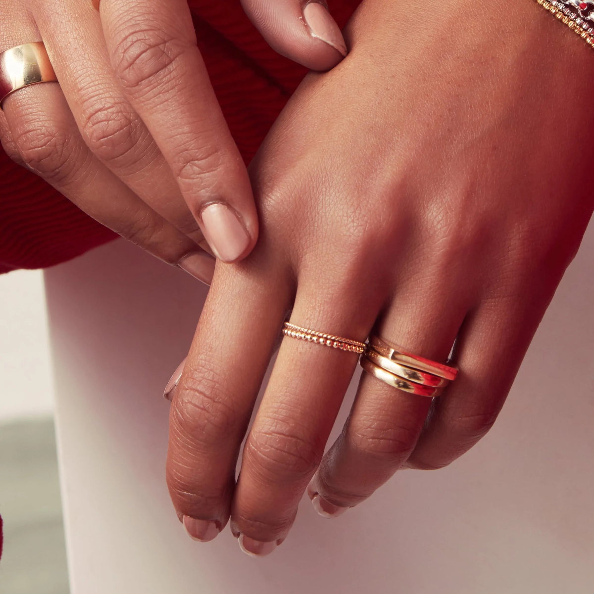 Close-up of hands wearing a Delicate Beaded Band, Delicate Twist Band, Thick Band, Geo Stacking Ring and two Tube Rings, all in yellow gold, against a light background. 