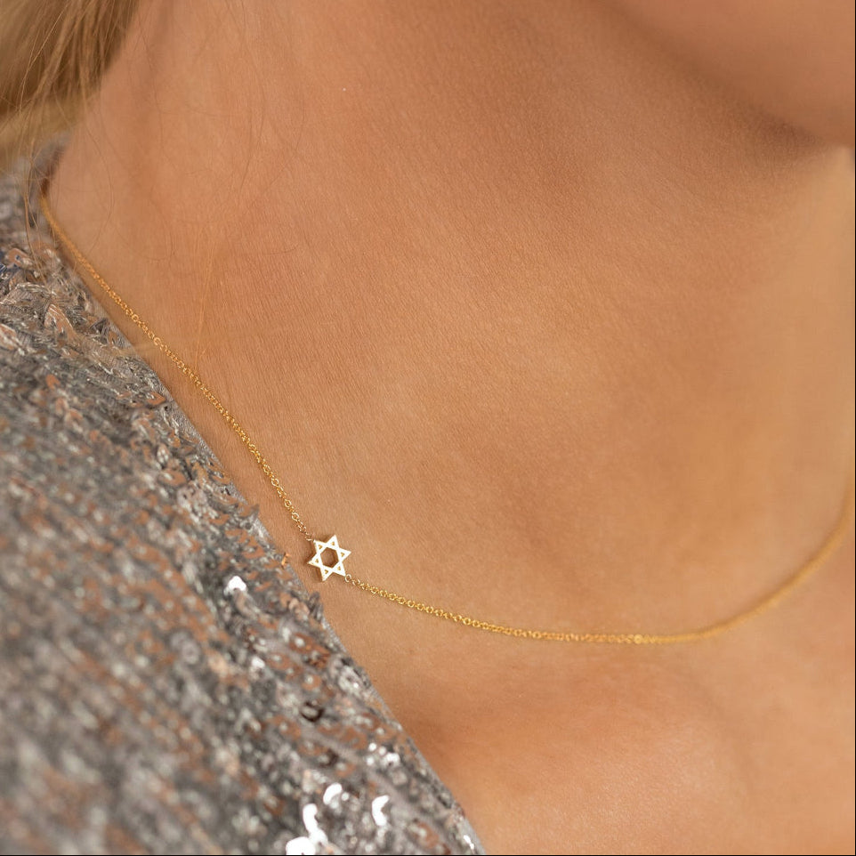 A close-up of a woman's neck wearing a 14k Gold Asymmetrical Charm Necklace - Star of David in yellow gold with a cable chain, along with Diamond Cascade Earrings, styled with a silver sequined top.