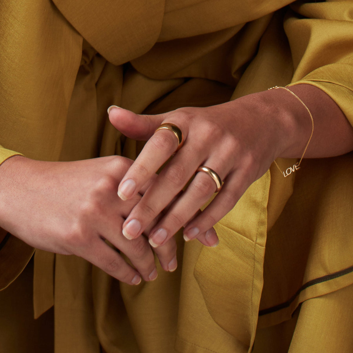A pair of hands wearing a yellow gold Thick Band, Tube Ring and Love Bracelet, with the person wearing mustard yellow clothing. 