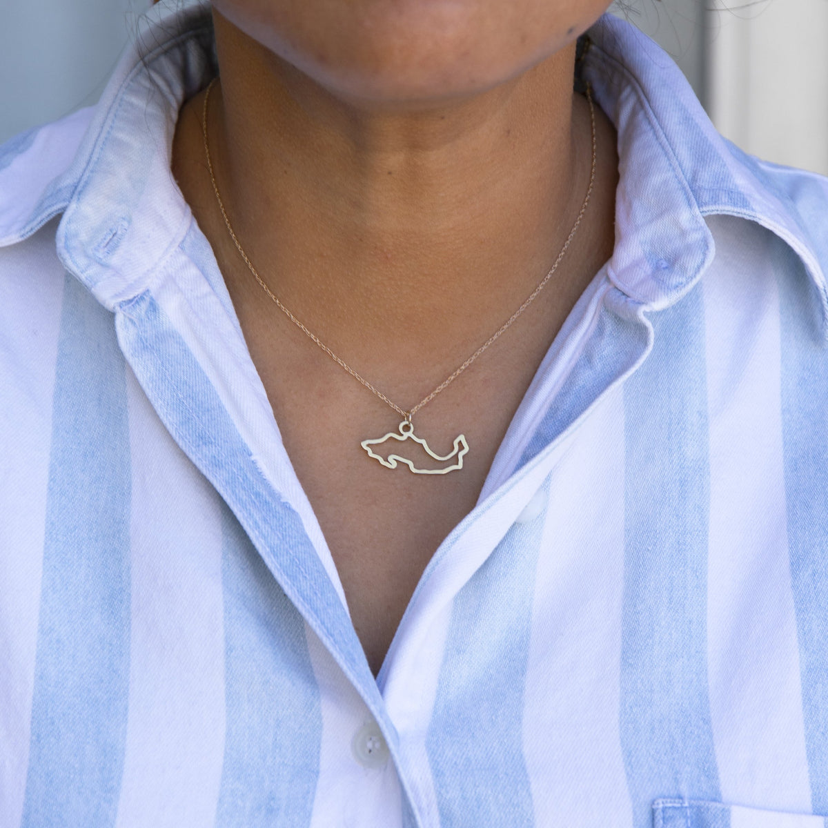 Close-up of a person's neck wearing a Mexico necklace in white gold with a blue and white striped shirt against a light grey blurred background.
