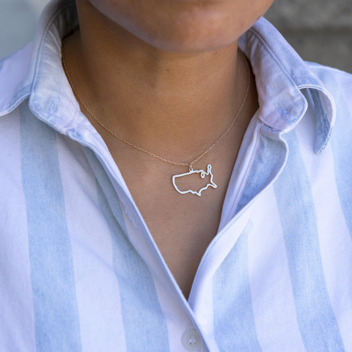 Close-up of a person's neck wearing a USA necklace in sterling silver with a blue and white striped shirt against a light grey blurred background.
