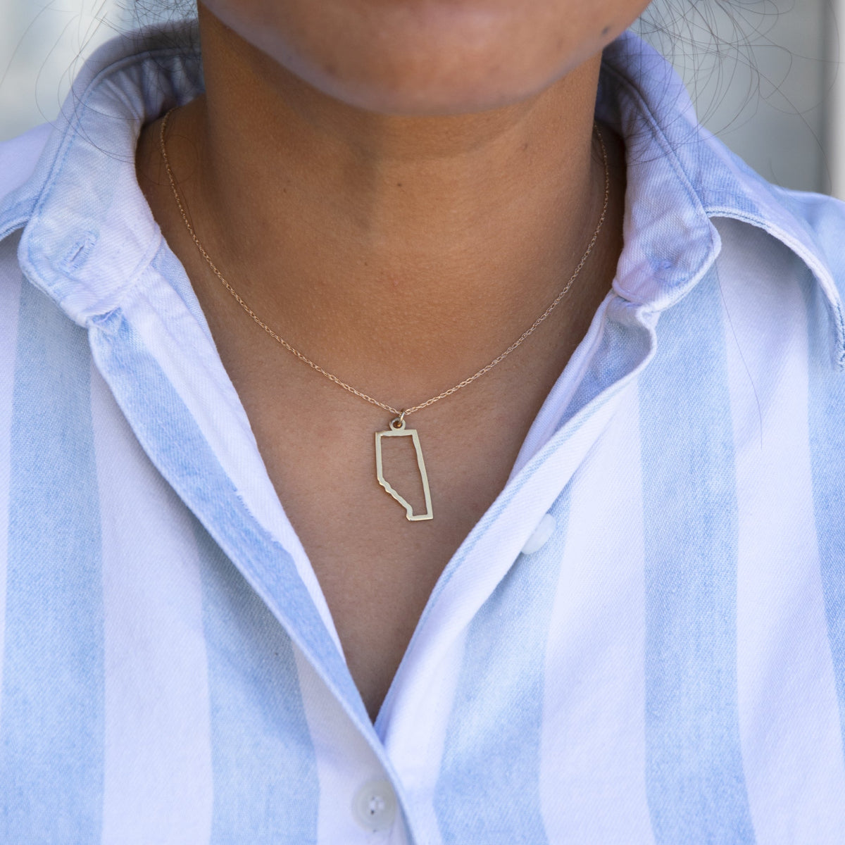 Close-up of a person's neck wearing an Alberta necklace with a blue and white striped shirt against a grey blurred background. 