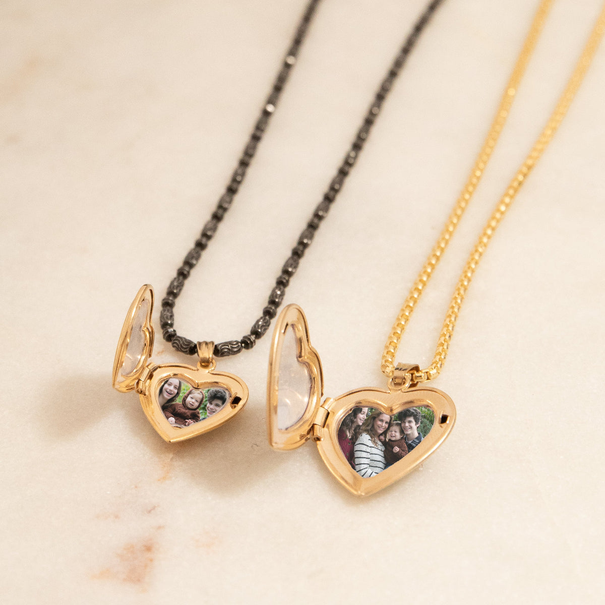 A Black Rhodium Antique Bead Chain with a Heart Locket Charm and a Box Chain with a Diamond Heart Locket Charm, both charms open with a family photo inside, against a cream background. 