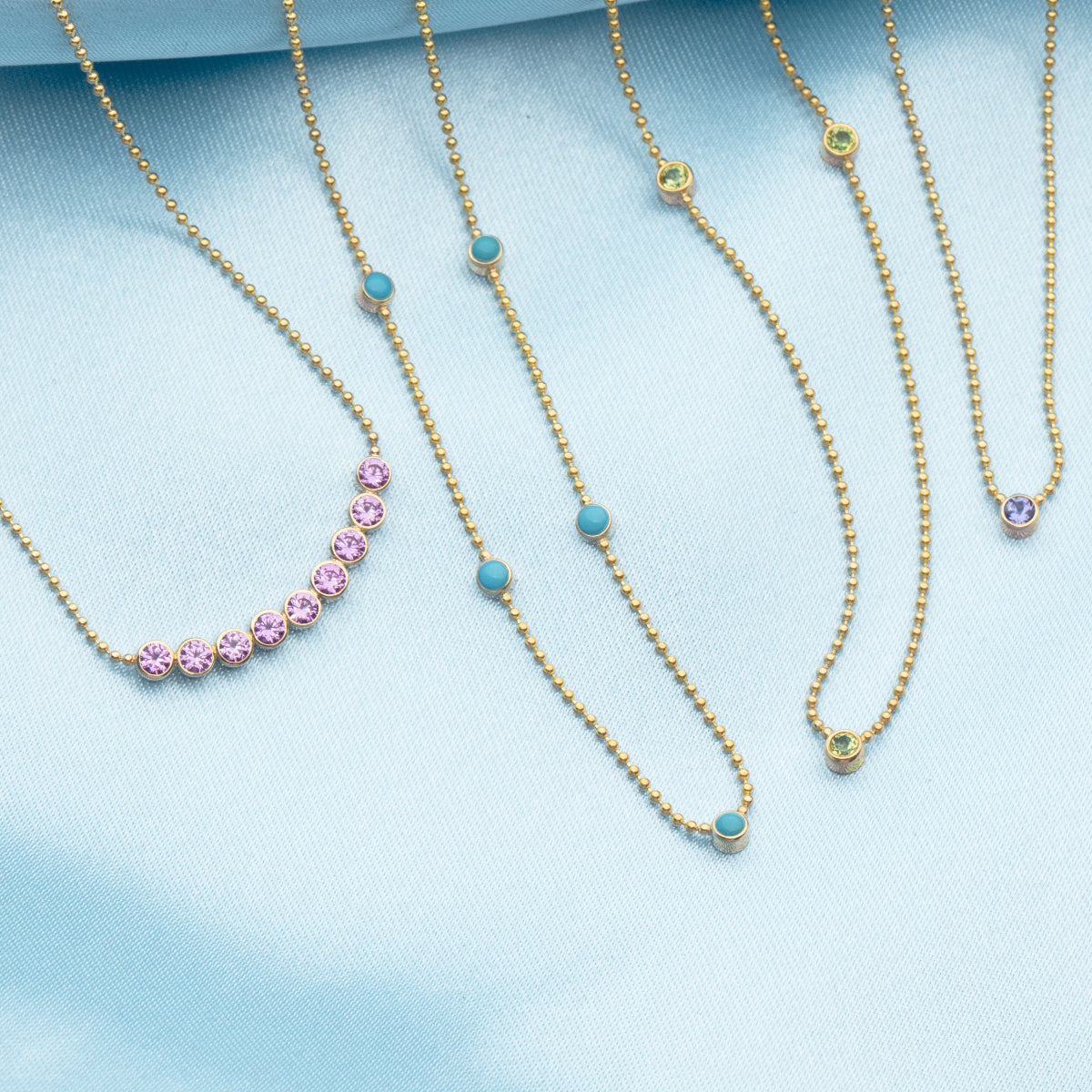 A Birthstone Arc Layering Necklace (Pink Tourmaline), Quintet Birthstone Layering Necklace (Turquoise), Triple Birthstone Layering Necklace (Peridot) and  Single Birthstone Layering Necklace (Tanzanite), all in yellow gold, against a light blue fabric background. 